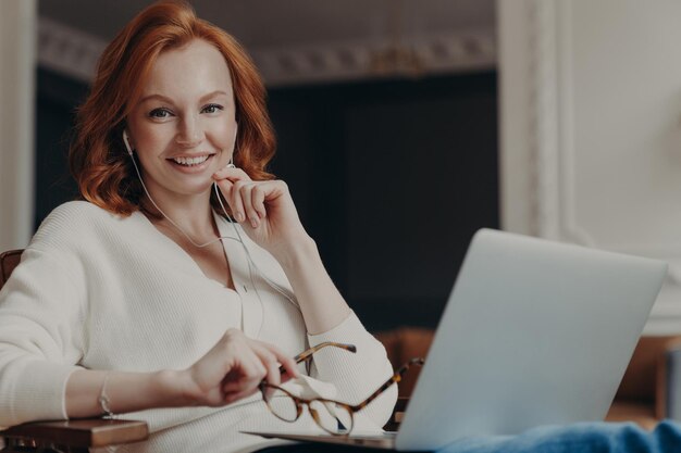 La mujer pelirroja positiva tiene una videollamada con un amigo a través de una computadora portátil y los auriculares disfrutan del tiempo para chatear a través de una cámara web tiene un seminario web en línea durante poses de trabajo remoto en el espacio de coworking