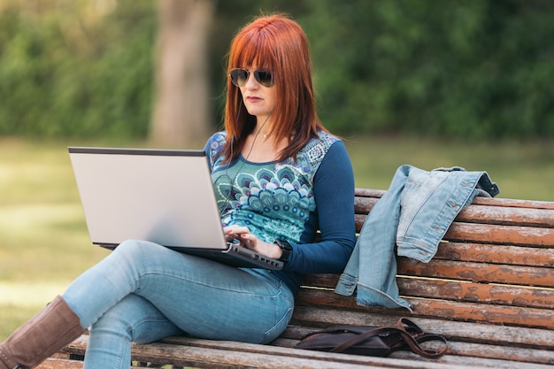 Mujer pelirroja sentada en un banco del parque usando una computadora portátil en un día soleado