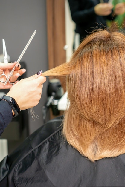 Mujer pelirroja sentada frente al espejo y recibiendo un corte de pelo largo rojo por parte de una peluquera en una peluquería vista trasera