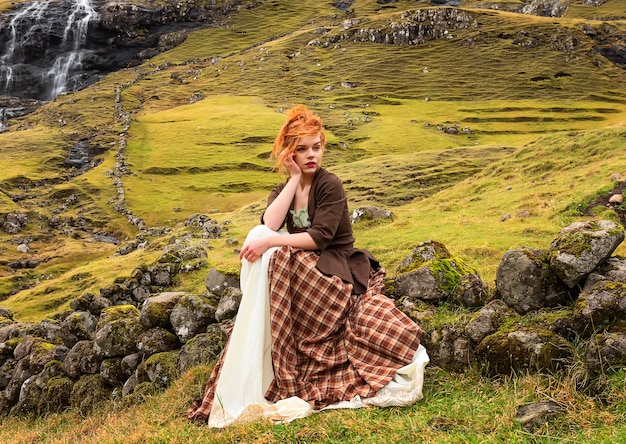 Una mujer pelirroja sentada sobre una valla de piedra con ropa pasada de moda Saksun Streymoy Faroe Island