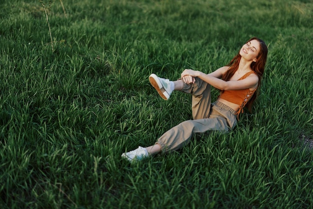 La mujer pelirroja se sienta en el parque sobre la hierba verde con un top naranja, pantalones verdes y zapatillas de deporte, y mira la puesta de sol de verano