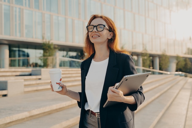 Mujer pelirroja soñadora en ropa formal lleva taza de café desechable de tableta de Bloc de notas