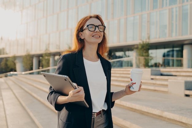 Mujer pelirroja soñadora en ropa formal lleva taza de café desechable de tableta de Bloc de notas