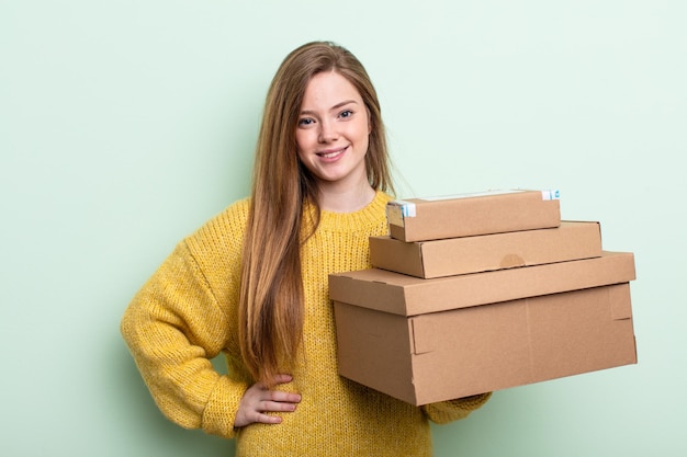 Mujer pelirroja sonriendo alegremente con una mano en la cadera y confiada. concepto de cajas de paquetes