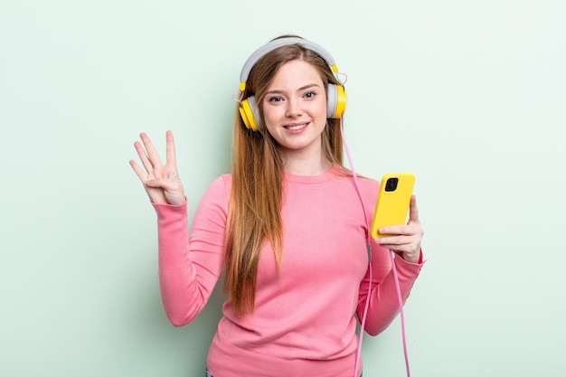 Mujer pelirroja sonriendo y luciendo amigable, mostrando el número cuatro. auriculares y concepto de teléfono inteligente