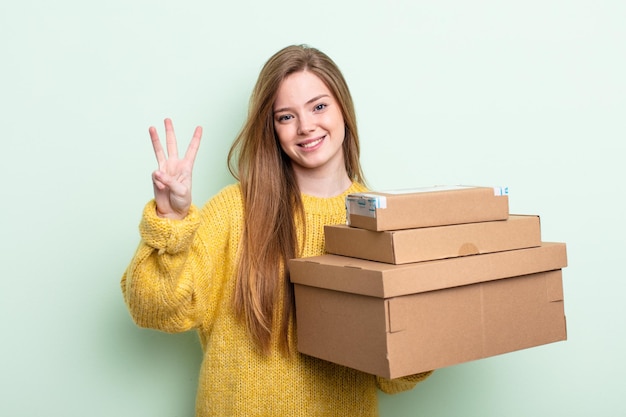 Mujer pelirroja sonriendo y luciendo amigable, mostrando el número tres. concepto de cajas de paquetes