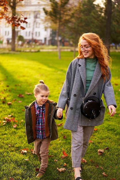 Mujer pelirroja sonriente y niño pequeño caminando sobre césped verde en el parque de la ciudad en un cálido día de otoño