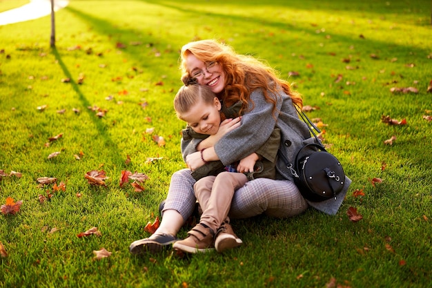 Mujer pelirroja sonriente y niño pequeño sentados en el césped en el parque de la ciudad en un cálido día de otoño joven mamá