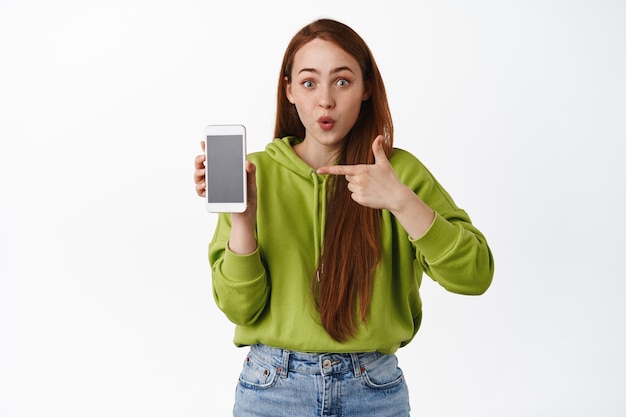 Foto mujer pelirroja sorprendida apuntando con el dedo a la aplicación de teléfono inteligente, jadeando de asombro, mostrando una aplicación interesante en el teléfono móvil en blanco