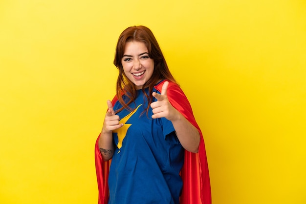 Mujer pelirroja de superhéroe aislada sobre fondo amarillo apuntando hacia el frente y sonriendo