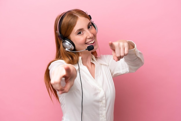 Mujer pelirroja de telemercadeo que trabaja con un auricular aislado en un fondo rosado te señala con el dedo mientras sonríe