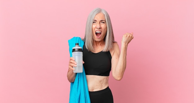 Mujer de pelo blanco de mediana edad gritando agresivamente con una expresión de enojo con una toalla y una botella de agua. concepto de fitness