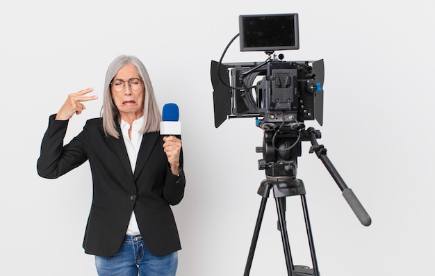 Mujer de pelo blanco de mediana edad que parece infeliz y estresada, gesto de suicidio haciendo un signo de pistola y sosteniendo un micrófono. concepto de presentador de televisión