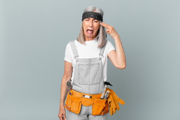 Mujer de pelo blanco de mediana edad que parece infeliz y estresada, gesto de suicidio haciendo un signo de pistola y usando ropa de trabajo y herramientas. concepto de limpieza