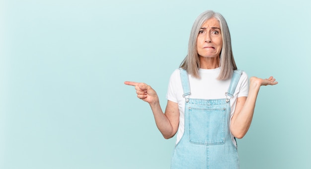 Mujer de pelo blanco de mediana edad que se siente desconcertada y confundida y dudando y apuntando hacia un lado