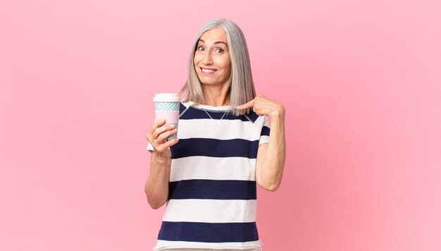 Mujer de pelo blanco de mediana edad que se siente feliz y apuntando a sí misma con un emocionado y sosteniendo un recipiente de café para llevar