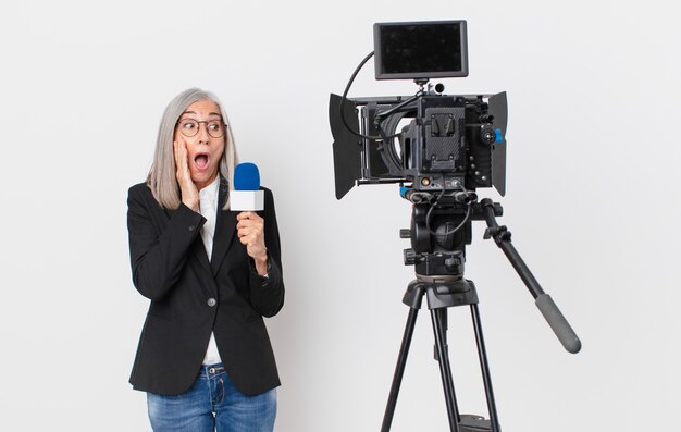 Mujer de pelo blanco de mediana edad que se siente feliz, emocionada y sorprendida y sosteniendo un micrófono. concepto de presentador de televisión