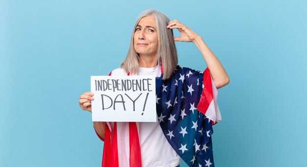 Mujer de pelo blanco de mediana edad que se siente perpleja y confundida, rascándose la cabeza. concepto del día de la independencia