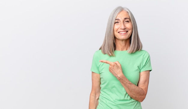 Foto mujer de pelo blanco de mediana edad sonriendo alegremente, sintiéndose feliz y apuntando hacia un lado y apuntando hacia el lado