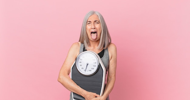 Mujer de pelo blanco de mediana edad sosteniendo una báscula. concepto de fitness y dieta