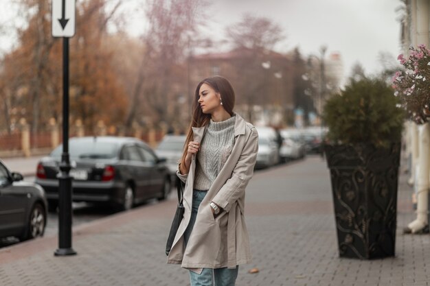 Mujer de pelo castaño en ropa de primavera y zapatillas de deporte con una bolsa de cuero posando a lo largo de la calle