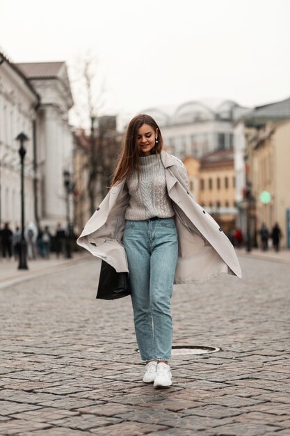 Mujer de pelo castaño en ropa de primavera y zapatillas de deporte con una bolsa de cuero posando a lo largo de la calle