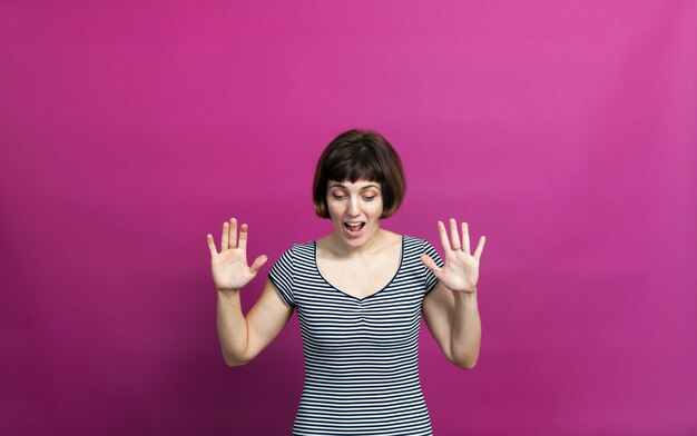 Mujer con pelo corto levantando las manos y mirando hacia abajo con sorpresa sobre fondo rosa