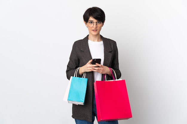 Mujer con pelo corto sobre un espacio aislado sosteniendo bolsas de la compra y escribiendo un mensaje con su teléfono celular a un amigo