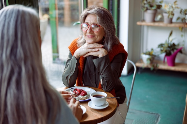 Mujer de pelo gris con gafas mira a un amigo sentado en una mesa pequeña en un acogedor café