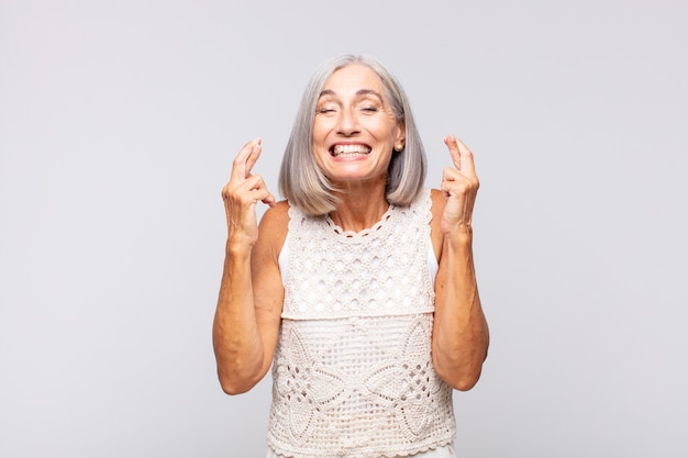 Mujer de pelo gris que se siente nerviosa y esperanzada, cruzando los dedos, rezando y esperando buena suerte