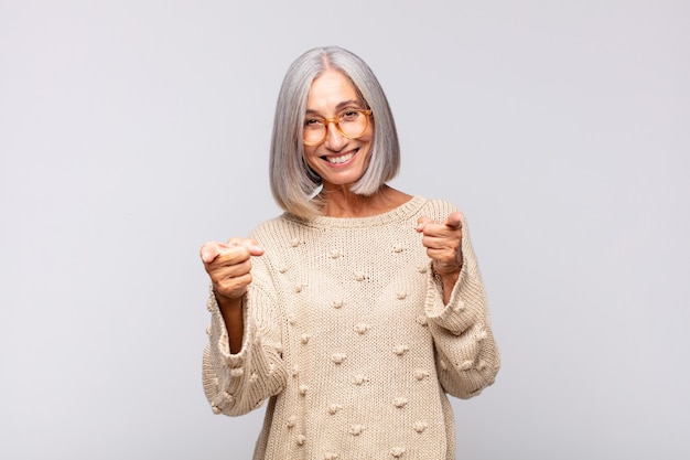 Mujer de pelo gris sonriendo con una actitud positiva, exitosa y feliz apuntando al frente, haciendo un signo de pistola con las manos