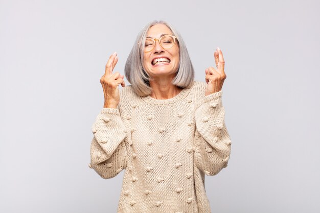 Mujer de pelo gris sonriendo y cruzando ambos dedos ansiosamente, sintiéndose preocupada y deseando o esperando buena suerte