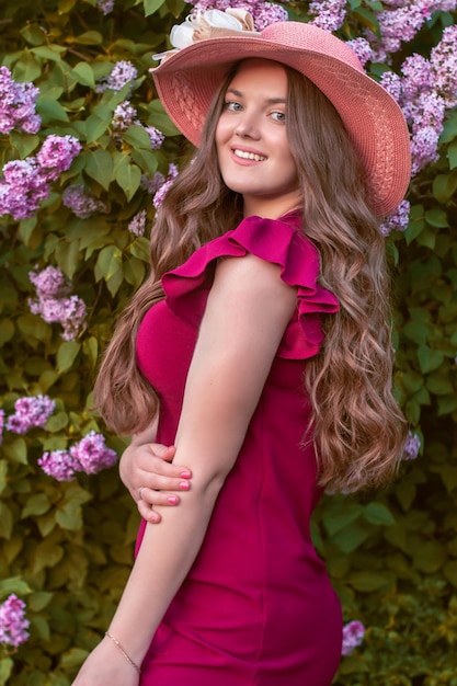 mujer de pelo largo y cabello castaño con un vestido morado se alza contra el fondo de un arbusto lila