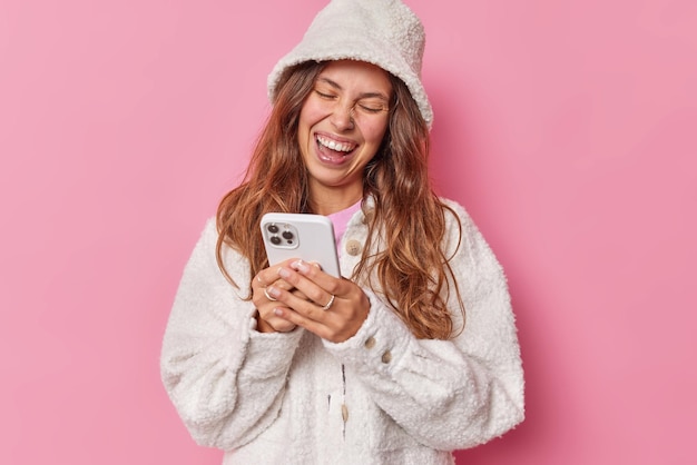 Mujer de pelo largo llena de alegría usa teléfonos móviles, mira videos divertidos, usa sombrero de piel y chaqueta, ve fotos después de la fiesta aislada sobre fondo rosa. Concepto de tecnología de emociones positivas de personas