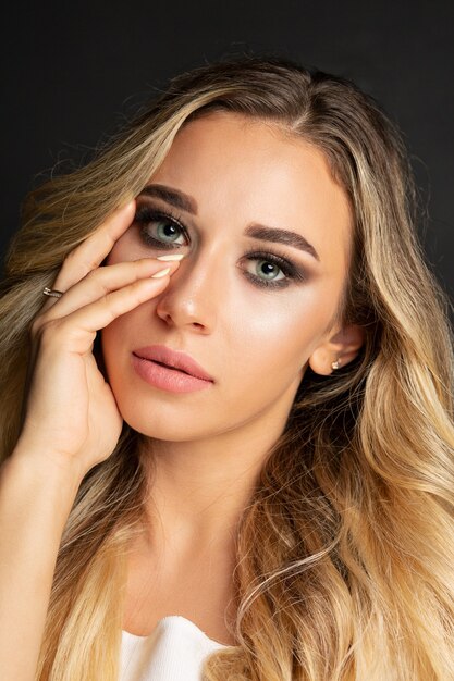 Mujer de pelo largo rubia glamorosa con maquillaje brillante posando en fondo negro de estudio