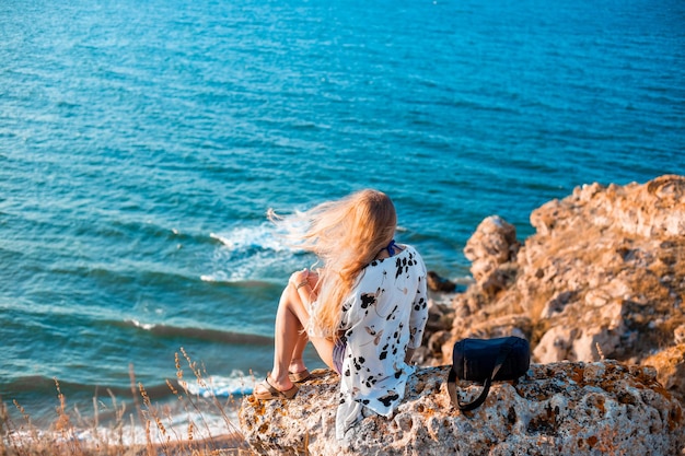 Una mujer con el pelo largo y rubio volando en el viento se sienta en la cima de una montaña y mira el paisaje marino