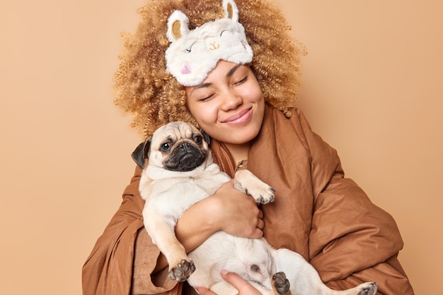 Una mujer de pelo rizado complacida sonríe agradablemente lleva un perro pug que va a dormir con una mascota envuelta en una pose de edredón en el dormitorio aislado sobre un fondo beige Animales domésticos y tiempo para dormir