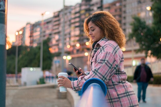 Mujer de pelo rizado con curvas mira su teléfono mientras bebe café en la calle