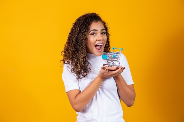 Mujer de pelo rizado emocionada en concepto de compras. Mujer joven con un carro en miniatura. Comercio electrónico y negocios. Carrito de compras. Mujer compradora. Fondo amarillo.