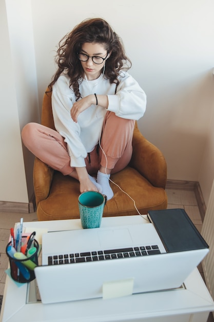 Mujer de pelo rizado con gafas está teniendo lecciones en línea en casa usando una computadora portátil y bebiendo un té