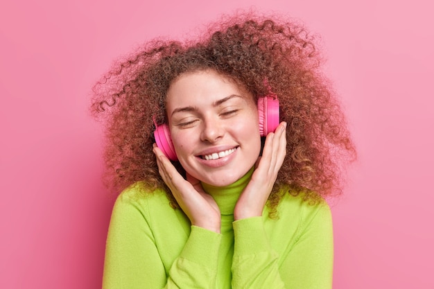 La mujer de pelo rizado hermosa relajada mantiene las manos en los auriculares estéreo cierra los ojos disfruta de la música favorita sonríe suavemente vestida con ropa casual aislada sobre la pared rosa. Estilo de vida juvenil