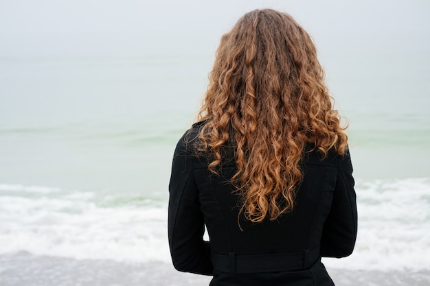 Mujer con el pelo rizado marrón en una capa negra que mira el mar en un día nublado