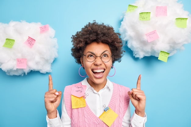 La mujer de pelo rizado positivo señala los dedos índices en las nubes con pegatinas de colores, lleva gafas redondas, muestra planes e información para recordar aislado sobre una pared azul.