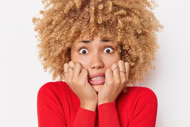 Una mujer de pelo rizado preocupada se ve ansiosa ante la cámara, aprieta los dientes y mira fijamente a la cámara, se siente nerviosa y asustada, usa un jersey rojo casual aislado sobre fondo blanco, ve una película de terror