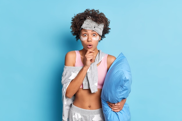 Mujer de pelo rizado sorprendida sostiene la barbilla y se ve sorprendida aplica parches de colágeno mira fijamente los ojos saltones sostiene la almohada se prepara para dormir aislado sobre una pared azul