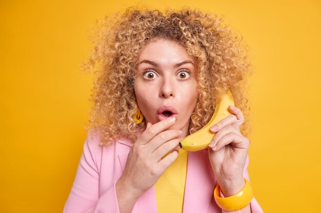 Mujer de pelo rizado sorprendida sostiene plátano maduro fresco cerca de la oreja finge llamar mantiene la boca abierta del asombro vestida con ropa formal posa contra la pared amarilla vívida. Reacción humana