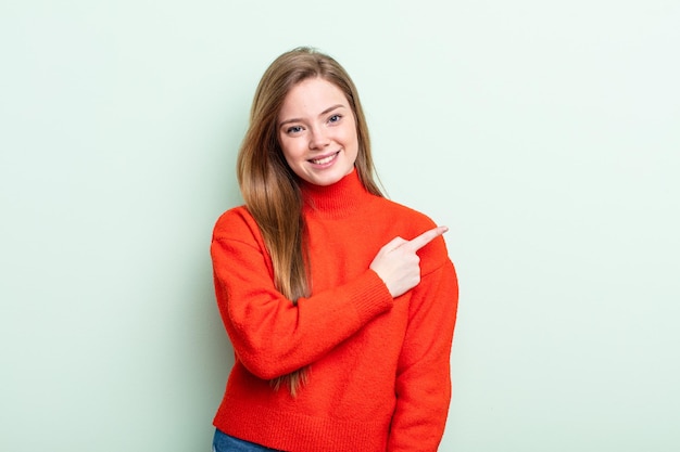 Mujer de pelo rojo caucásico sonriendo alegremente, sintiéndose feliz y apuntando hacia un lado y hacia arriba, mostrando el objeto en el espacio de la copia