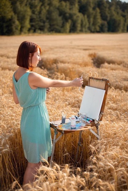 Mujer con pelo rojo corto en vestido azul comienza a pintar en el campo de tiro horizontal de trigo maduro