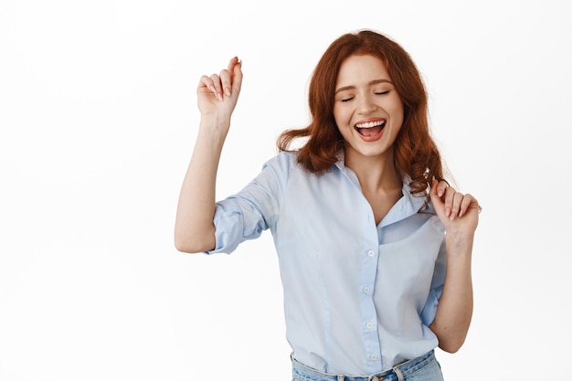 Mujer con pelo rojo rizado, divirtiéndose en el tiempo libre, sonriendo despreocupada y feliz, disfrutando del ocio, de pie en blusa en blanco