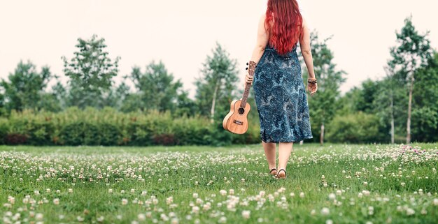 Mujer con el pelo rojo con un ukelele en sus manos, caminando sobre la hierba verde en el parque en un día de verano. Imagen panorámica con lugar para texto.